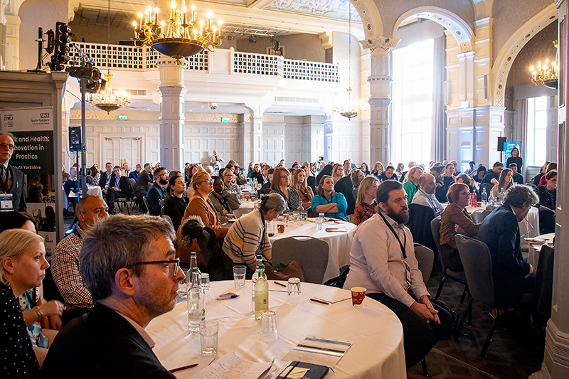 An event in a grand old building. People are sat round tables and are looking towards the front of the room