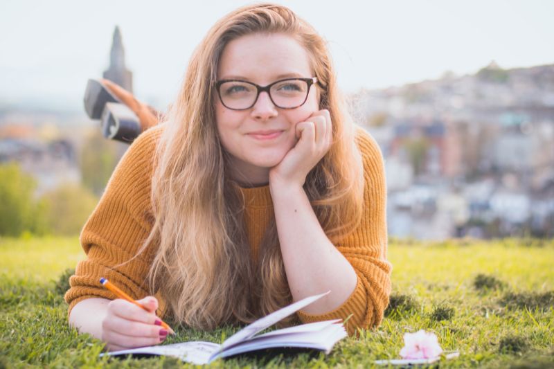 Girl laying on her front on the grass, leaning on her elbows and writing in a notebook.