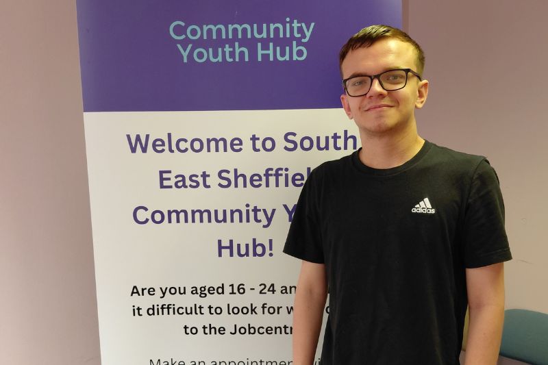 Young man wearing glasses stood in front of a purple and white banner that says: Community Youth Hub.