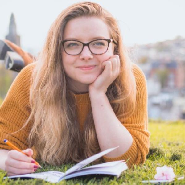 Girl laying on her front on the grass, leaning on her elbows and writing in a notebook.